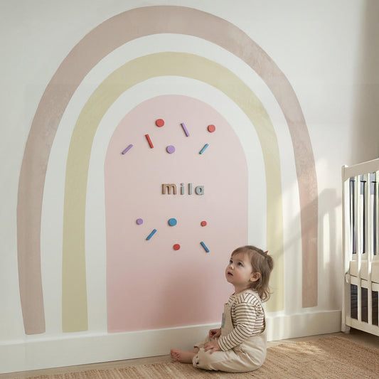 Child sitting in a room with a rainbow wall decoration and name 'mila'.