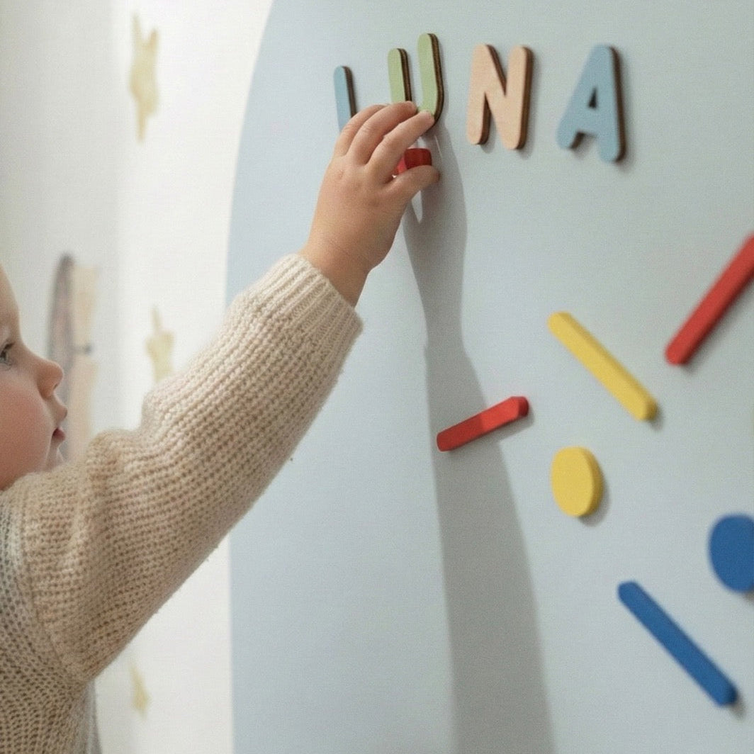 Close-up of a toddler placing wooden name magnets on a blue magnetic arch wall in a children’s room
