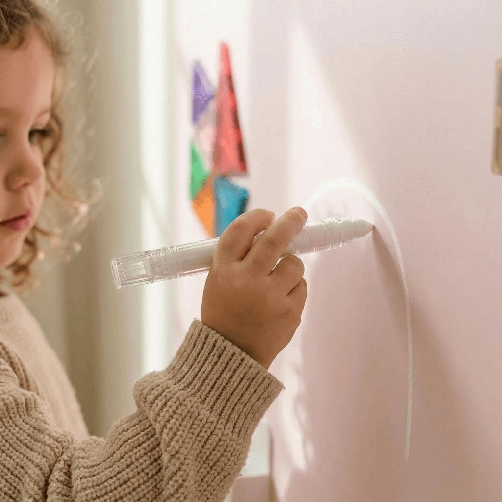 Child holding a white marker writing on a light-colored wall magnetic wall