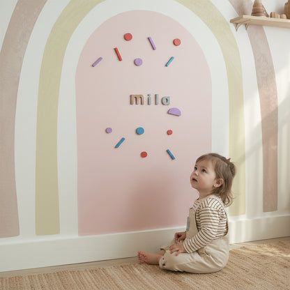 Toddler sitting in front of a pastel rainbow magnetic arch wall designed for creative play