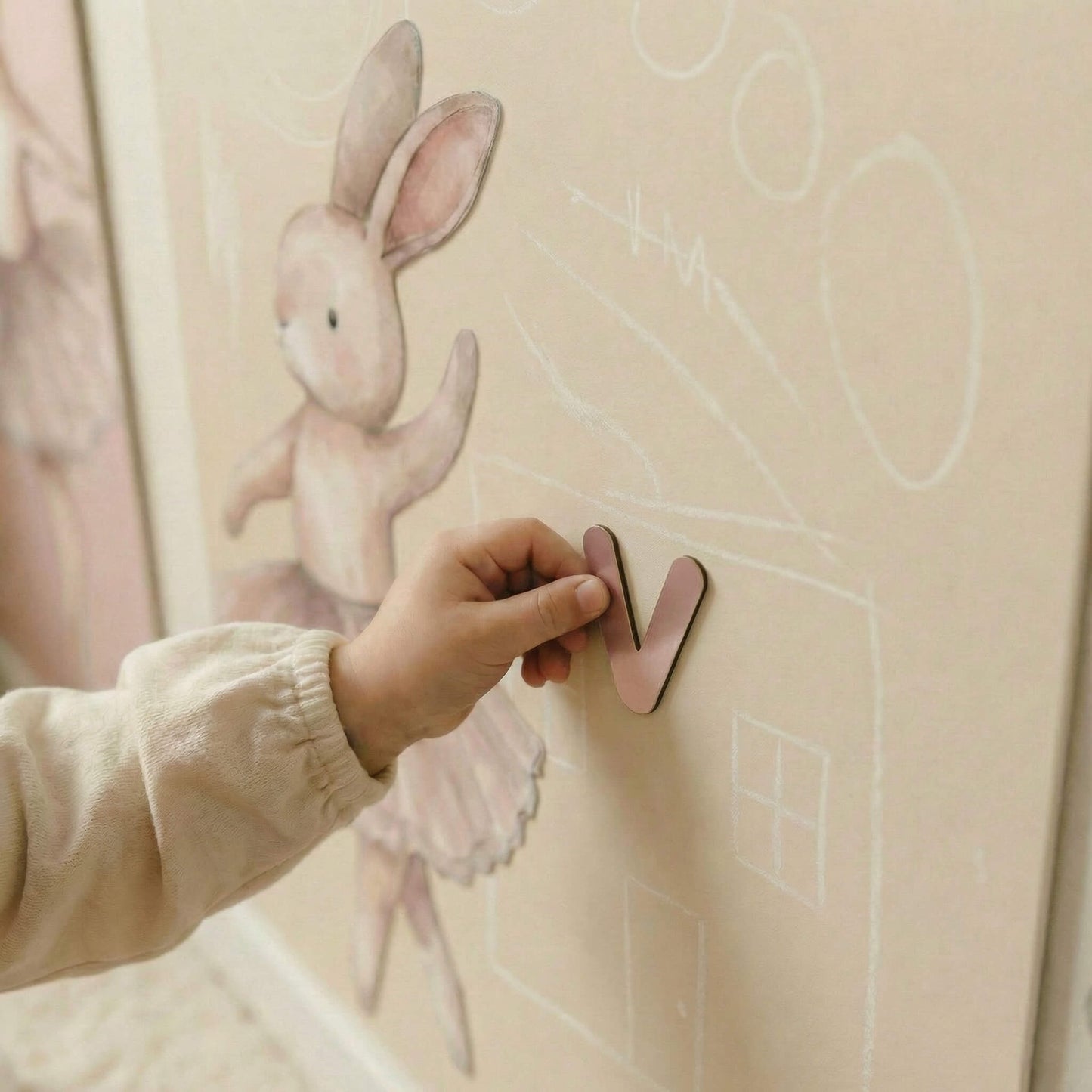 Child's hand placing a pink letter 'V' on a wall with a cartoon rabbit and house drawing.