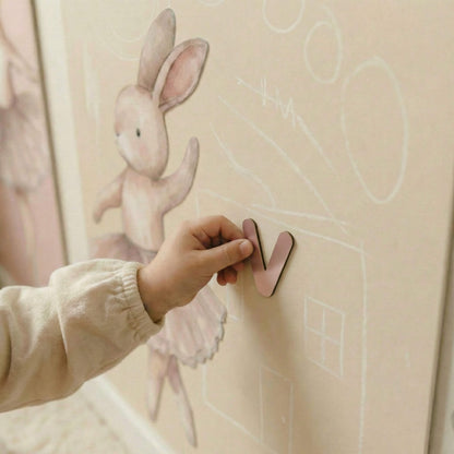 Child's hand placing a pink letter 'V' on a wall with a cartoon rabbit and house drawing.