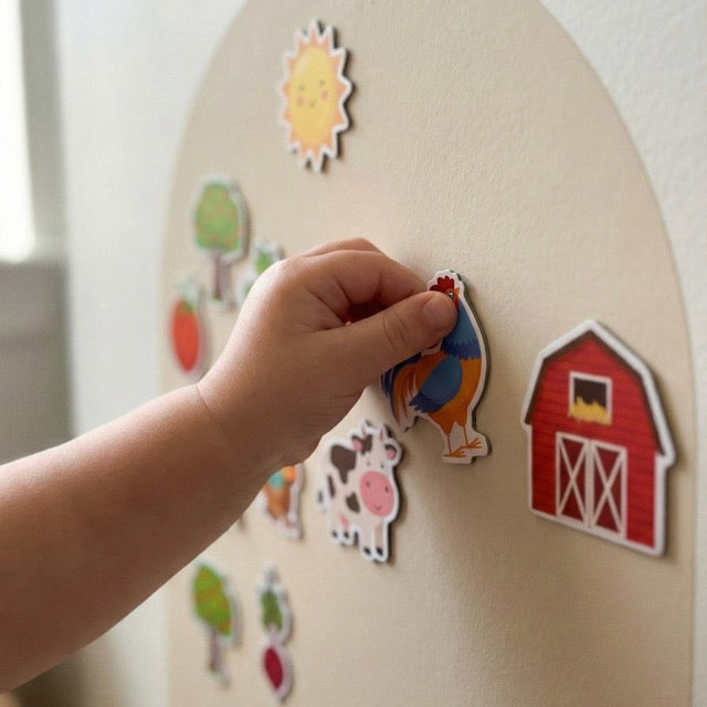 A child's hand placing a rooster magnet onto a beige magnetic arch wall play panel.